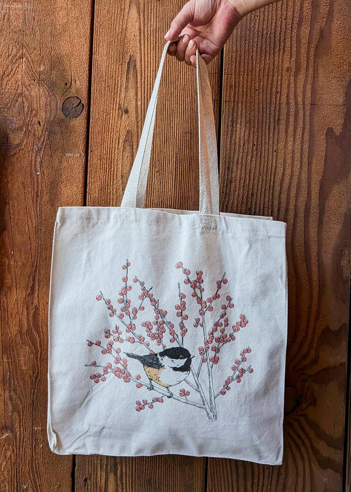 Light canvas tote bag held by handles, featuring chickadee with red berry branch print