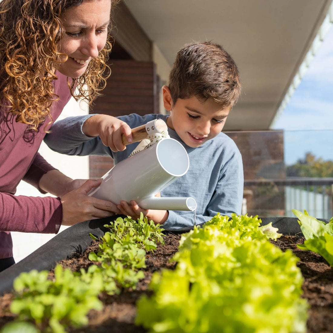 A mother and child watering leafy greens on a sunny balcony garden using container planters
