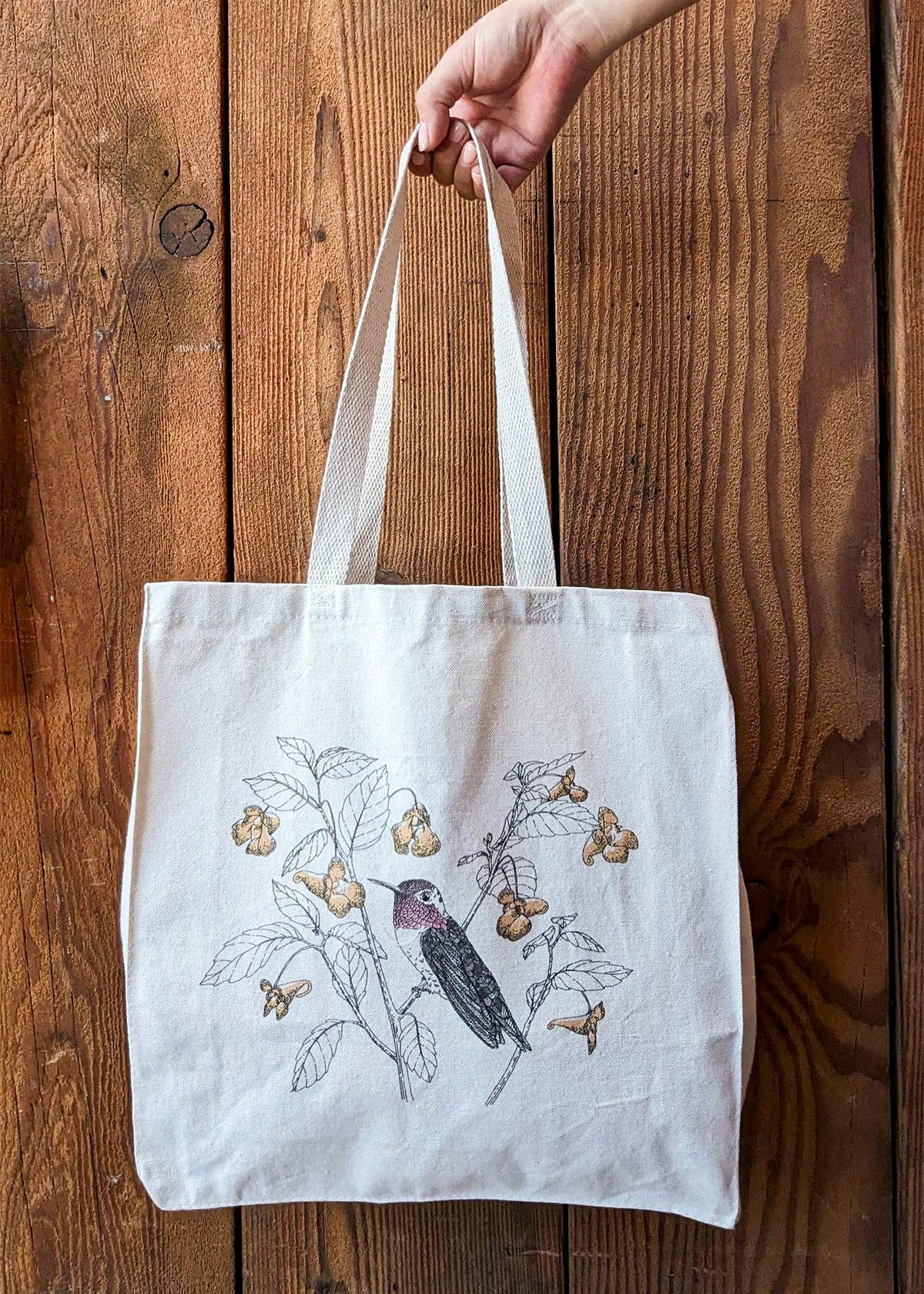 Off-white canvas tote bag in hand, hummingbird and wildflower letterpress print
